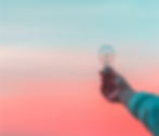 Hand holding a lightbulb against a soft pastel sunset sky, blending pink and blue hues, creating a tranquil and contemplative mood.