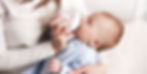 Woman in a white sweater feeding a baby with a bottle. Baby wears a blue polka-dot outfit, in a soft, light-filled setting. Calm mood.