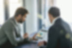Two men in suits discuss a bar chart on a tablet in a modern office. Bright natural light fills the room, creating a focused mood.