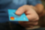 Hand holding a blue debit card with visible "Debit" text, against a blurred background. The focus is on the card being presented.