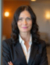 Woman with long curly hair in a black blazer and white shirt smiles confidently with arms crossed; warm, blurred indoor background.