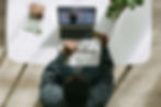 Person at a white desk shops online on a laptop, holding a credit card. Items include a wallet, money, and a potted plant. Bright setting.