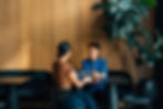 Man in a blue shirt and woman with a tablet talk in a wood-paneled cafe. Green plant in the background, warm lighting, focused mood.