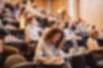 Students in a lecture hall; a woman in the foreground writes in a notebook. Wooden walls create a studious atmosphere.