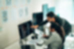 Two people in an office work on code at a computer. Papers with diagrams are pinned on the wall. Bright lighting and focused mood.