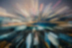 Blurred cityscape with colorful light streaks radiating from center, creating a dynamic, energetic effect against a dark evening sky.